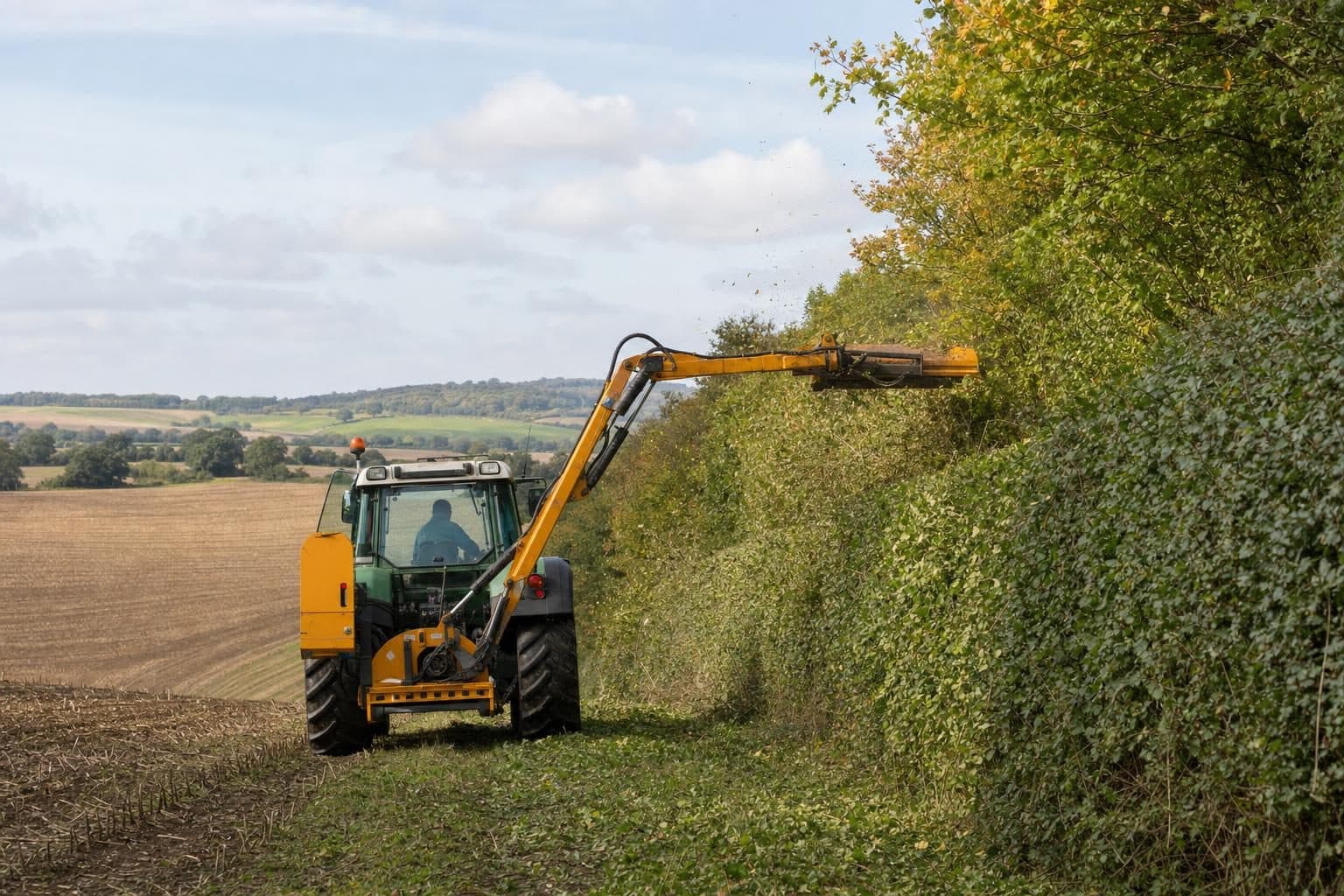 A tractor with a flail trimming a farm hedgerow beside a field